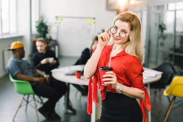 Cheerful young beautiful blond woman in glasses and red shirt standing at her working place on office with cup of coffee. Multiracial young creative people in modern office.