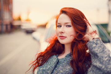 Red-haired Model in a coat posing against the backdrop of the sunset on the street.