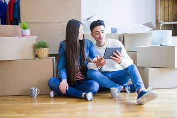 Young asian couple sitting on the floor of new apartment arround cardboard boxes, using touchpad tablet and smiling at new home