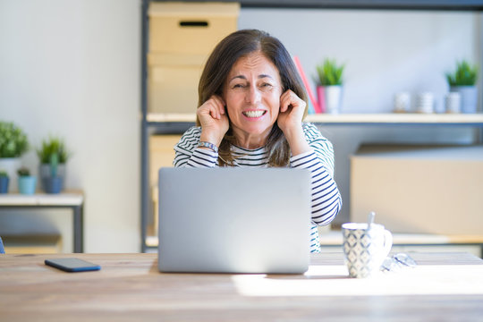 Middle Age Senior Woman Sitting At The Table At Home Working Using Computer Laptop Covering Ears With Fingers With Annoyed Expression For The Noise Of Loud Music. Deaf Concept.