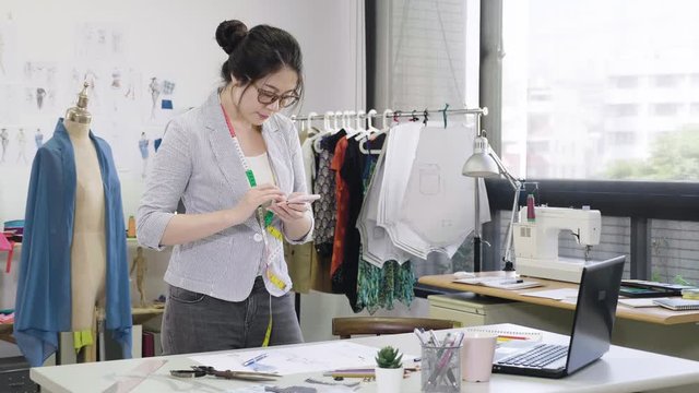 Beautiful asian woman fashion designer taking photos of sketches lying on table by smart phone. professional girl worker making creative flat lays. studio light full of sewing items tools equipment