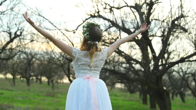 Ballerina in white wedding dress poses with spread out hands in apple orchard among trees in ballerinas outdoors on grass during spring day closeup view captured in 120 FPS