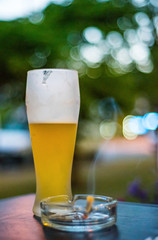 glass of beer on a wooden table with ashtray with smoke cigarette