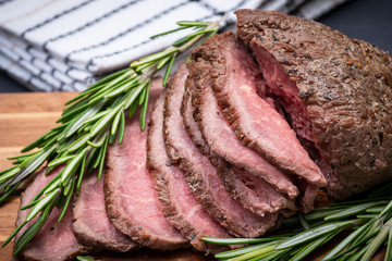 Sliced Grass Fed juicy Corn Roast Beef garnished with Rosemary Fresh Herb on natural wood cutting board.