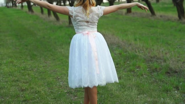 Ballerina in white wedding dress poses on tiptoe legs with spread arms in apple orchard among trees in ballerinas outdoors on grass during spring day closeup view captured in 120 FPS