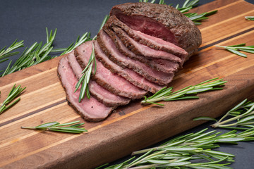 Sliced Grass Fed juicy Corn Roast Beef garnished with Rosemary Fresh Herb on natural wood cutting board.