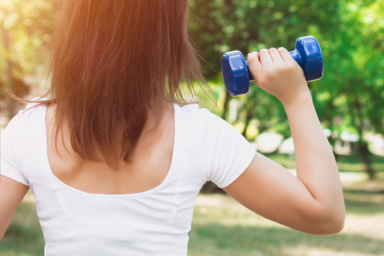 Sport training concept. Girl in a white T-shirt stands with her back and holds a blue dumbbell on the background of the park. The girl is engaged in fitness in nature