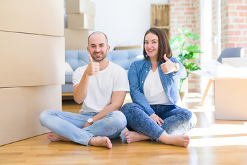 Young couple sitting on the floor arround cardboard boxes moving to a new house doing happy thumbs up gesture with hand. Approving expression looking at the camera with showing success.