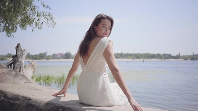 Portrait beautiful young girl with long brunette hair wearing a long white summer fashion dress sitting on the waterfront. Leisure pretty woman looking at the camera outdoors.