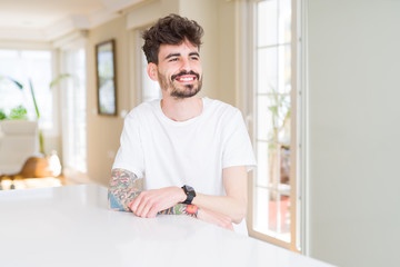 Young man wearing casual t-shirt sitting on white table looking away to side with smile on face, natural expression. Laughing confident.