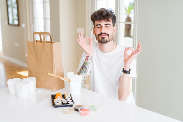 Young man eating asian sushi from home delivery relax and smiling with eyes closed doing meditation gesture with fingers. Yoga concept.