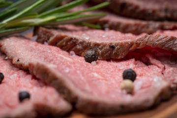 Sliced Grass Fed juicy Corn Roast Beef garnished with Fresh Rosemary Herb and Rainbow Peppercorns on natural wood cutting board.