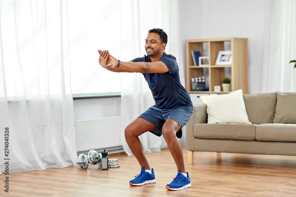 Wall mural sport and healthy lifestyle concept - smiling indian man with fitness tracker exercising and doing squats at home