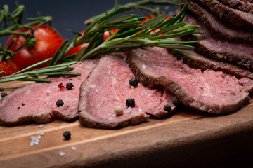 Sliced Grass Fed juicy Corn Roast Beef garnished with Tomatoes, Fresh Rosemary Herb and Rainbow Peppercorns on natural wooden cutting board.