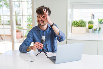 Young man recording podcast using microphone and laptop with happy face smiling doing ok sign with hand on eye looking through fingers