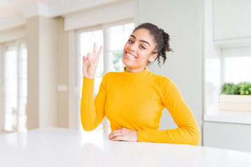 Beautiful african american woman with afro hair wearing a casual yellow sweater smiling looking to the camera showing fingers doing victory sign. Number two.