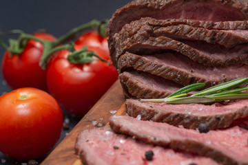 Sliced Grass Fed juicy Corn Roast Beef garnished with Tomatoes, Fresh Rosemary Herb and Rainbow Peppercorns on natural wooden cutting board.