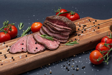 Sliced Grass Fed juicy Corn Roast Beef garnished with Tomatoes, Fresh Rosemary Herb and Rainbow Peppercorns on natural wooden cutting board.