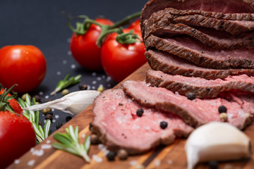 Sliced Grass Fed juicy Corn Roast Beef garnished with Tomatoes, Fresh Rosemary Herb, Garlic and Rainbow Peppercorns on natural wooden cutting board.