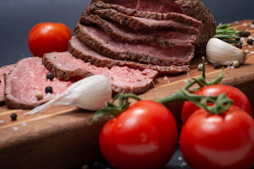 Sliced Grass Fed juicy Corn Roast Beef garnished with Tomatoes, Fresh Rosemary Herb, Garlic and Rainbow Peppercorns on natural wooden cutting board.