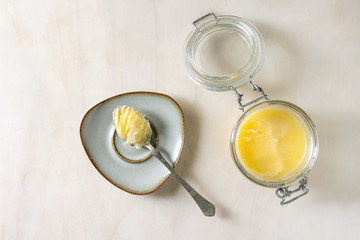 Homemade Melted ghee clarified butter in open glass jar and spoon on saucer over white marble background. Flat lay, space - obrazy, fototapety, plakaty