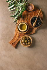 Variety of green and black whole olives in olive oil served in bowls with fresh baked ciabatta bread, pink salt and young olive wood branches over brown texture background. Flat lay, copy space
