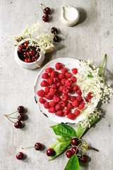 Fresh cherry and raspberry berries in ceramic mug and plate, elderflowers, jug of cream over gray texture background. Flat lay, space