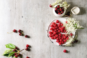 Fresh cherry and raspberry berries in ceramic mug and plate, elderflowers, jug of cream over gray texture background. Flat lay, space