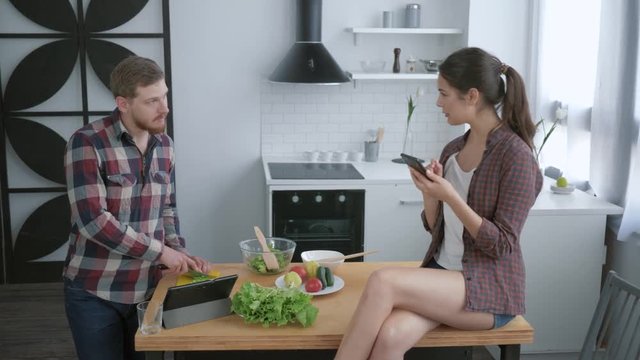 weight normalization, man prepares healthy vegetarian salad on breakfast and woman sitting on kitchen table with smartphone in hands and takes selfie photo at cuisine