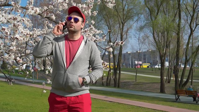 Man in sunglasses talks on smartphone in city park. Man in red baseball cap with red smartphone