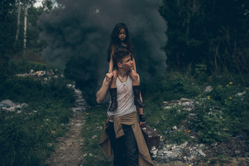 Homeless father and daughter walk through the garbage dump on background of smoke.