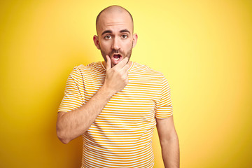 Young bald man with beard wearing casual striped t-shirt over yellow isolated background Looking fascinated with disbelief, surprise and amazed expression with hands on chin