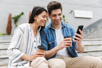 Image of cheery couple drinking takeaway coffee and using smartphone on city stairs outdoors