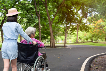 Asian senior woman in wheelchair with child girl supporting disabled grandparent on walking green nature,grandmother and granddaughter enjoy on street  in summer at park,elderly care,family concept