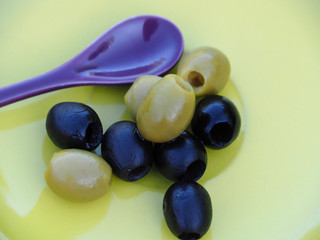 Green and blue olives with colored spoon on yellow plate background. Delicious fruit close-up.