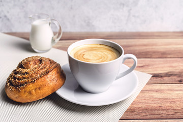 Perfect breakfast of a homemade cinnamon bun, coffee and glass milk jug on wooden table. Rustic style. Close-up.
