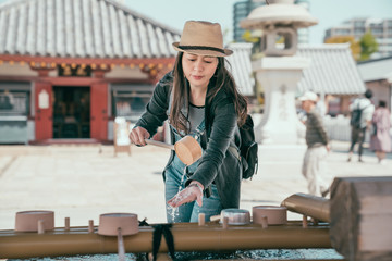 Japanese purification fountain in shitennoji shrine. korean woman tourist wear hat washing hands before entering temple. young girl traveler with backpack using bamboo ladle with clear water.