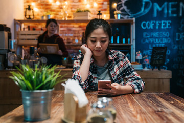 Young asian woman texting message on mobile phone waiting for beer at bar in dark night sitting alone. girl waitress staff standing in counter preparing making meal for customer. lady use cellphone