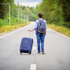 Fototapeta na wymiar Female tourist with a travel bag on wheels and a backpack walks away on the road