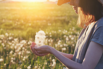 pretty young girl wearing linen dress holding glass jar full of beautiful fluffy white fresh fragile dandelion flowers, saving happy moments, best summer memories, sunny morning background, copy space