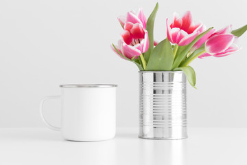 Enamel mug mockup with pink tulips in a pot on a white table.
