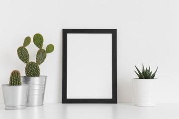 Black frame mockup with various types of cactus and a succulent plant on a white table. Portrait orientation.