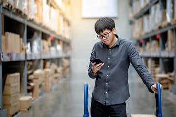 Asian guy shopper standing with shopping cart in warehouse inventory aisle checking shopping list on smartphone. Buying or purchasing factory goods. Shopaholic concept