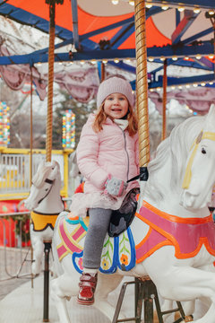 Adorable smiling Caucasian child girl riding on merry go round carousel horse at Christmas winter market outdoor. Happy child having fun celebrating New Year.