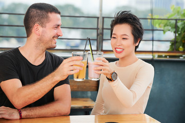 Happy young couple sitting at the table and toasting with glasses of fresh juice during their meeting in cafe