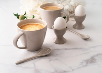 Boiled eggs in ceramic egg cups, two cups of coffee with ceramic spoons on background of beautiful white peony. Breakfast concept. Copy space.