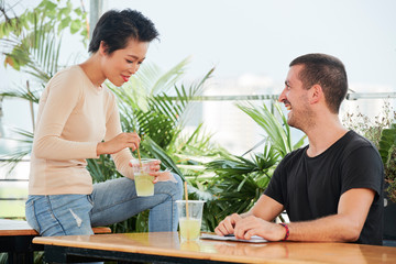 Young multiethnic couple sitting at the table drinking lemonade talking to each other and have a funny time together in cafe