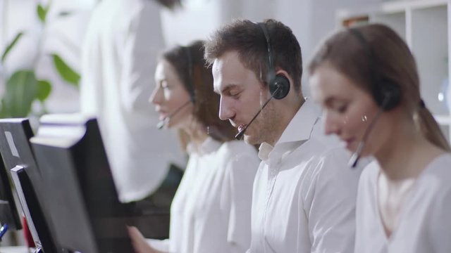 Business and technology concept - helpline operator with headphones in call centre working hard, then looks straight to camera and smiles happily. Politeness, good mood, positive emotions. Female