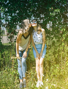 Two hipster girls are standing near the ivy leaves fence outdoors. Funny happy emotions of sisters. Best friends photo session.Stylish and fashionable women. Travel lifestyle concept.