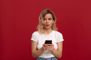 Portrait of a girl with curly blond hair in a white t-shirt on a red background. Surprised model in wireless headset shows screen of a mobile phone.